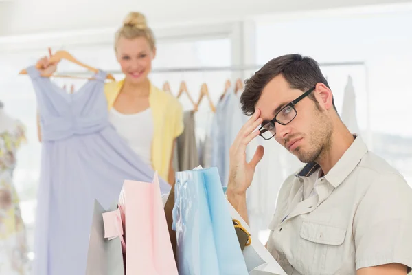 Bored man with shopping bags while woman at clothes rack — Stock Photo, Image
