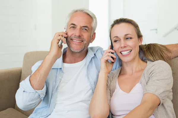 Pareja feliz sentada en el sofá hablando en sus teléfonos —  Fotos de Stock