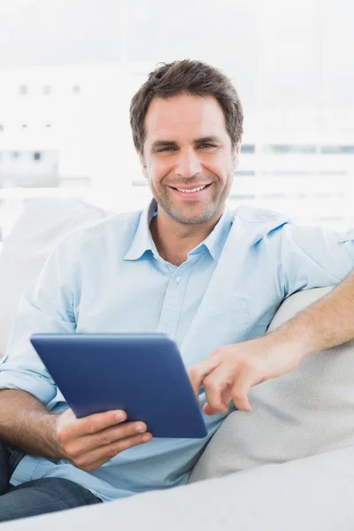 Красивый улыбающийся мужчина сидит на диване, используя свой планшетный компьютер — стоковое фото