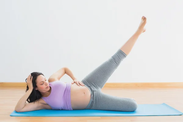 Lächelnde schwangere Brünette, die auf Matte liegt und ihr Bein hebt — Stockfoto