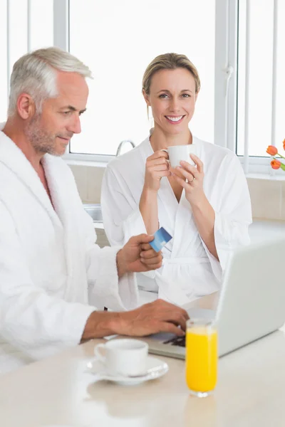 Smiling couple using laptop at breakfast in bathrobes — Stock Photo, Image