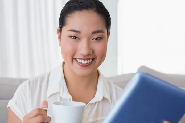 コーヒーを飲んでタブレット pc を使用してソファの上に座っている女性の笑みを浮かべてください。 — ストック写真