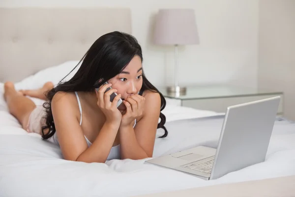 Pretty asian lying on bed talking on phone looking at laptop — Stock Photo, Image