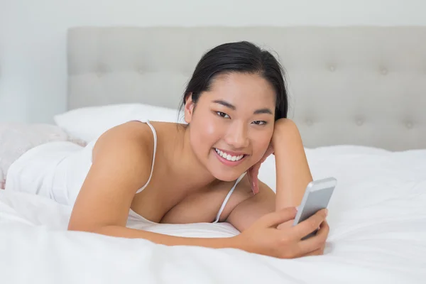 Lächelnde asiatische Frau, die auf dem Bett liegt und einen Text schickt — Stockfoto
