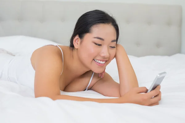 Улыбающаяся азиатка лежит на кровати и шлёт сообщение. — стоковое фото