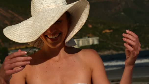 Rubia sonriente moviendo su sombrero de sol de su cara en la playa — Vídeos de Stock