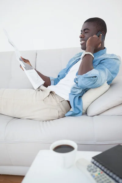 Casual homme couché sur le canapé sur le téléphone tenant journal — Photo