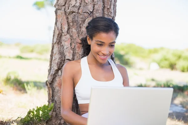 Fit mujer sentada contra el árbol usando el ordenador portátil — Foto de Stock