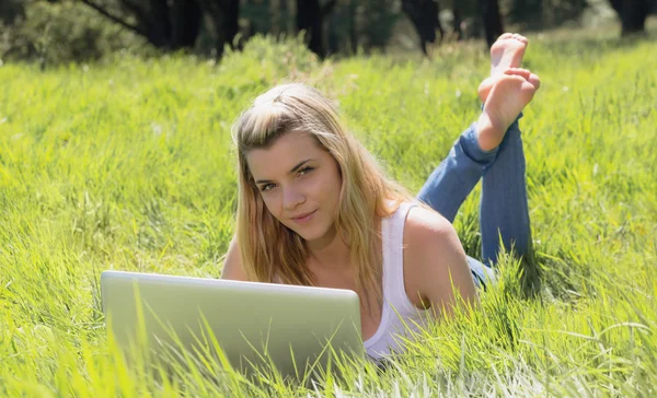 Bastante rubia acostada en la hierba usando el portátil sonriendo a la cámara — Foto de Stock