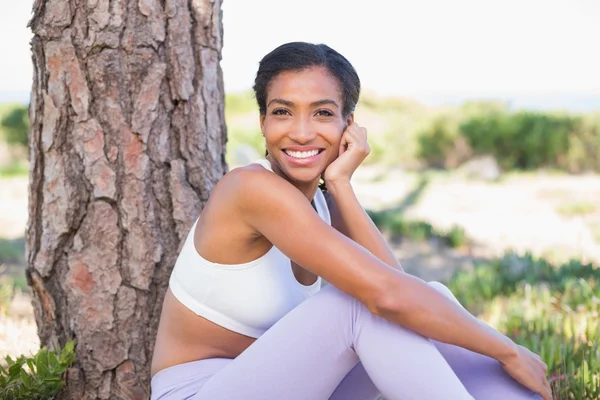 Fit woman sitting against tree smiling at camera — Stock Photo, Image