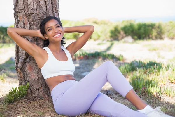 Fit mujer sentada contra el árbol sonriendo a la cámara — Foto de Stock