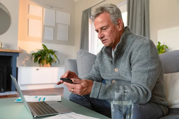 专注的高加索老人坐在沙发上 做文书工作 使用智能手机和笔记本电脑 积极的退休生活方式 — 图库照片
