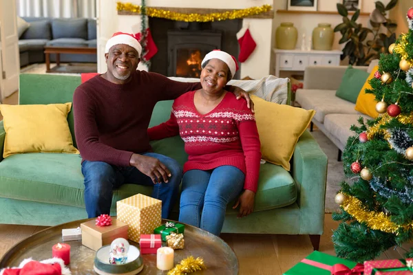 クリスマスの時にソファに座っているサンタの帽子の幸せなアフリカ系アメリカ人のシニアカップル 引退生活やクリスマスのお祭りを自宅で祝い — ストック写真