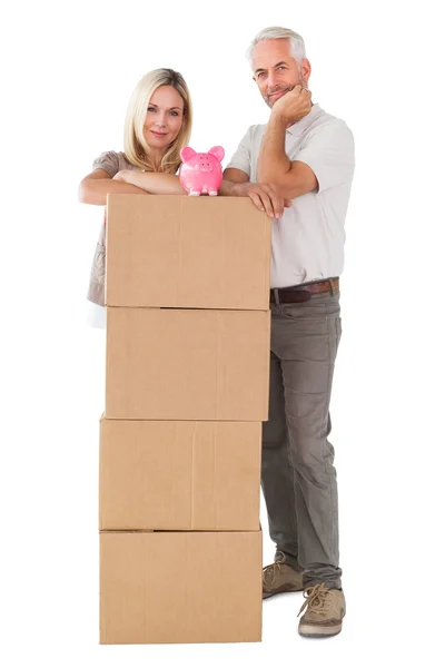 Happy couple leaning on pile of moving boxes with piggy bank — Stock Photo, Image