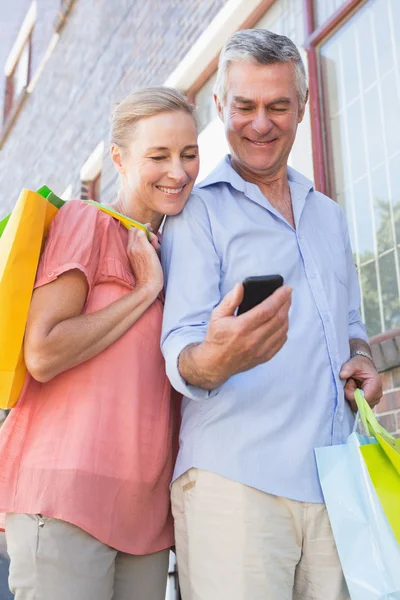 Happy senior couple looking at smartphone holding shopping bags — Stock Photo, Image