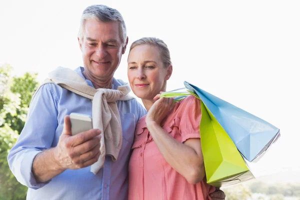 Happy senior couple looking at smartphone holding shopping bags — Stock Photo, Image