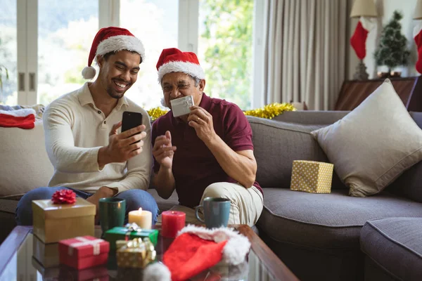 スマートフォンのクリスマスビデオ通話を作るサンタの帽子の幸せな出産の大人の息子と高齢者の父親 クリスマスやお祭りや通信技術は — ストック写真