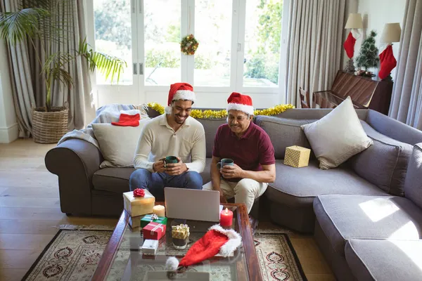 サンタの帽子の幸せな出産の大人の息子と高齢者の父親は ラップトップのクリスマスビデオ通話を行います クリスマスやお祭りや通信技術は — ストック写真