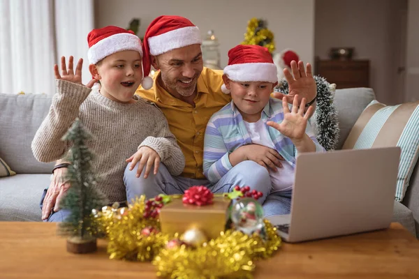 クリスマスの間に自宅のノートパソコンでビデオ通話をしている間に 白人の父と2人の息子が手を振っています クリスマスの時期に流行した19種のパンデミックの間の社会的距離は — ストック写真