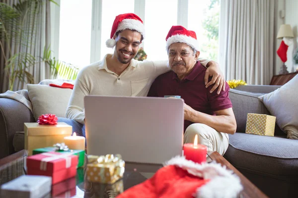 サンタの帽子の幸せな出産の大人の息子と高齢者の父親は ラップトップのクリスマスビデオ通話を行います クリスマスやお祭りや通信技術は — ストック写真