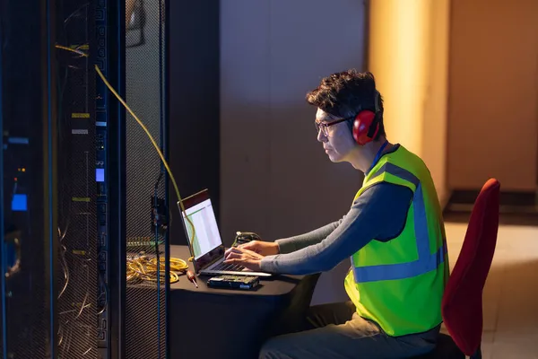 亚洲男性工程师带着耳塞 在电脑服务器室使用笔记本电脑 数据库服务器管理和维护概念 — 图库照片