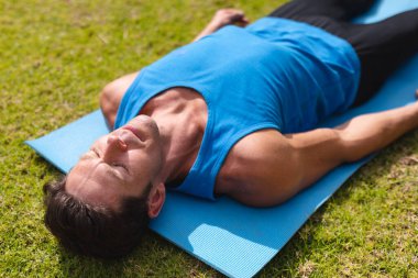 Young man in sportswear practicing shavasana on exercise mat in public park. yoga, healthy lifestyle and body care. clipart