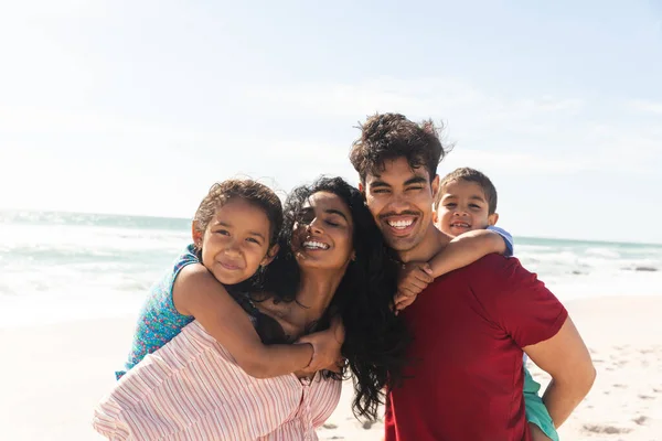 Happy biracial father and mother giving piggyback rides to kids at beach against sky with copy space. family, lifestyle and weekend.