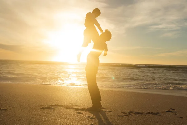 夕暮れ時には 海辺で息子を抱えて空を背景に照明を当てた父親の姿 家族生活や週末 — ストック写真