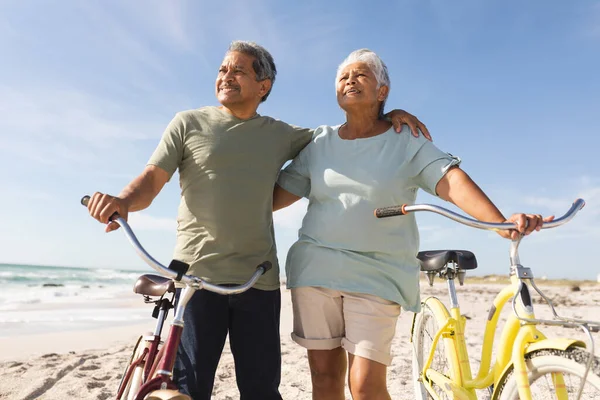 Uśmiechnięta Para Seniorów Odwracająca Wzrok Rowerami Plaży Przed Niebem Słoneczny — Zdjęcie stockowe