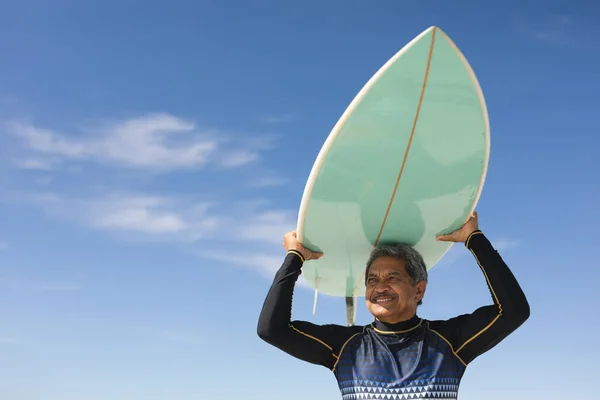 青い空に対して日当たりの良いビーチで頭の上にサーフボードを運ぶ出生シニア男性の低角度ビュー ウォータースポーツやアクティブライフスタイル — ストック写真