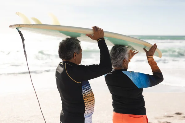 多人種のシニアカップルが一緒に日当たりの良いビーチで海岸沿いの頭の上にサーフボードを運ぶ歩いています ウォータースポーツやアクティブライフスタイル — ストック写真
