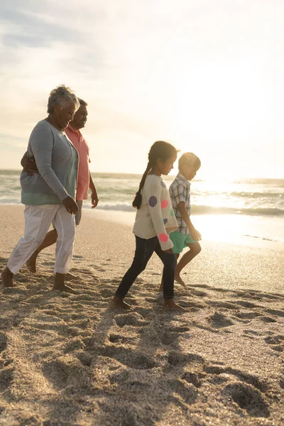 ビーチで孫の後ろを歩く腕を持つ多人種の祖父母の側のビュー — ストック写真