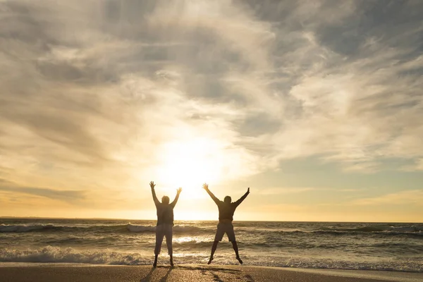 Carefree Multiracial Senior Couple Jumping Arms Raised Beach Sky Sunset — Stock Photo, Image