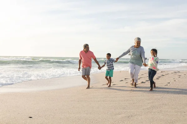 多人種の孫たちが祖父母と手を取り合い 空に向かってビーチを歩く — ストック写真