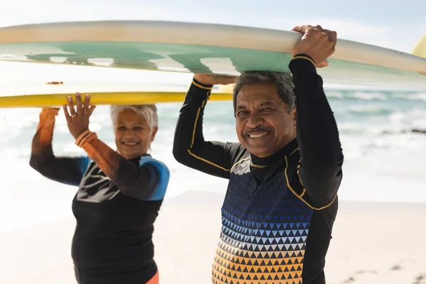 日当たりの良いビーチで頭の上にサーフボードを運ぶ多人種のシニア男性と女性の笑顔の肖像画 ウォータースポーツやアクティブライフスタイル — ストック写真