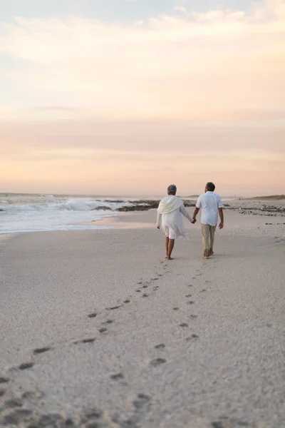 Wielorasowa Para Seniorów Spacerująca Zostawiając Ślady Stóp Plaży Podczas Zachodu — Zdjęcie stockowe