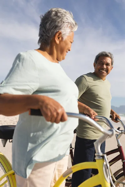 日当たりの良いビーチに自転車で立っている間 幸せな高齢者の多人種のカップルがお互いを見て 活動的な生活と交通機関 — ストック写真