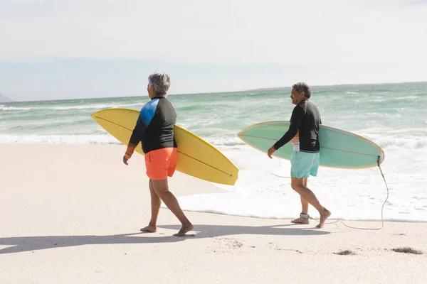 日当たりの良いビーチで海岸を歩いてサーフボードを運ぶ多人種のシニアカップルの完全な長さ ウォータースポーツやアクティブライフスタイル — ストック写真