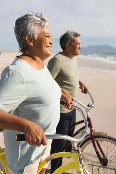 日当たりの良いビーチで海岸沿いの自転車で離れて見て笑顔の高齢者の多人種カップルの側面図 活動的な生活と交通機関 — ストック写真
