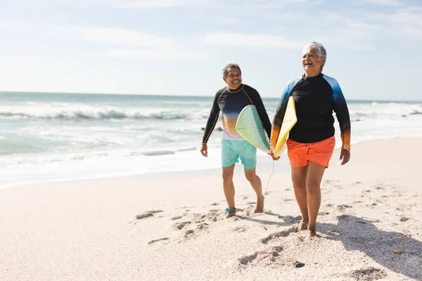 日当たりの良いビーチで砂の上にサーフボードで歩く幸せな多人種シニアカップルの完全な長さ ウォータースポーツやアクティブライフスタイル — ストック写真