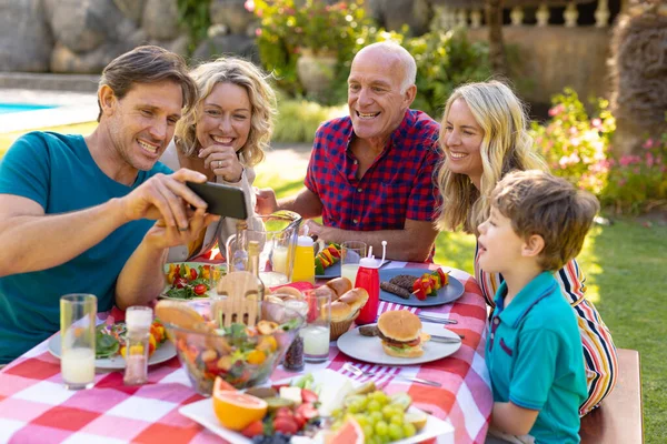白人の家族は庭のテーブルに座ってスマートフォンを共有しています 週末のライフスタイルの概念は — ストック写真