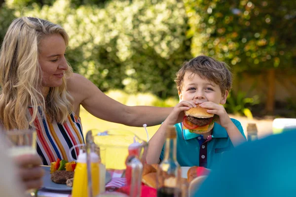 庭で家族と一緒にバーガーを食べる息子を見てブロンドの白人の母親 週末のライフスタイルの概念は — ストック写真