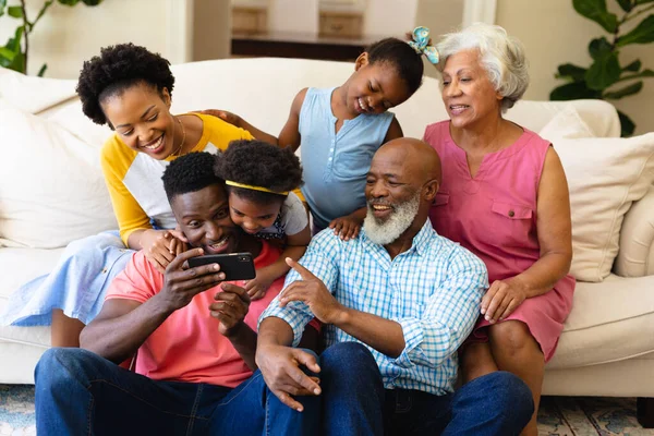 アフリカ系アメリカ人の3世代の家族が自宅のソファに一緒に座ってスマートフォンを使用しています 家族愛と団結の概念を変えずに — ストック写真