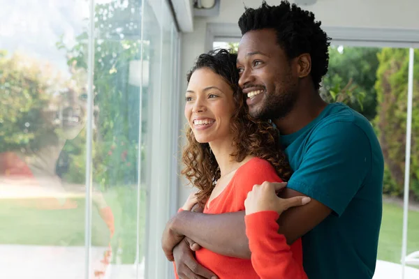 Jovem Casal Biracial Feliz Abraçando Enquanto Olha Pela Janela Casa — Fotografia de Stock