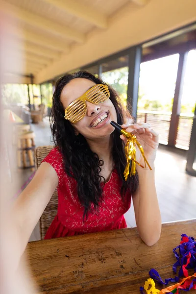 Cheerful biracial young woman with party horn and shutter shades enjoying party on video call. unaltered, video call, vitiligo and distant party celebration.