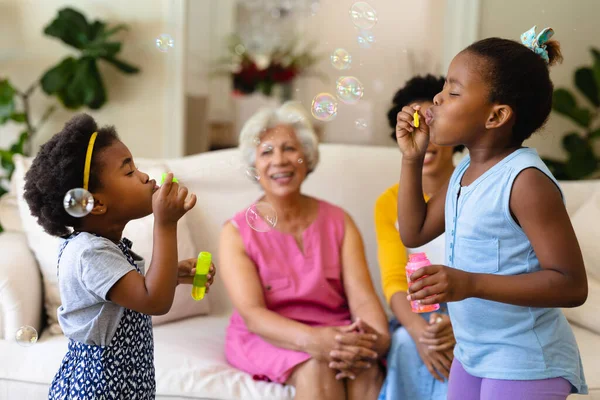 Dos Hermanas Afroamericanas Soplando Burbujas Mientras Abuela Madre Miran Sentadas —  Fotos de Stock