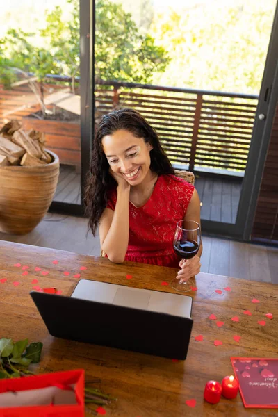Happy biracial young woman with red wine video calling through laptop at restaurant. unaltered, online dating, video call, wireless technology, vitiligo and distant valentine day celebration.