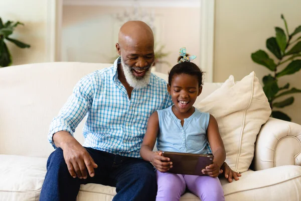 ソファに座っている間にデジタルタブレットを使用して孫娘とアフリカ系アメリカ人の先輩の笑顔 変化のない無線技術家族生活余暇活動国内生活の概念 — ストック写真