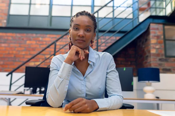 Portrait of confident african american young businesswoman with hand on chin at desk in office. unaltered, creative business, sitting and modern office concept.