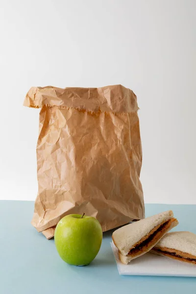 Green apple with paper bag and peanut butter and jelly sandwich on table against gray background. unaltered, healthy food, copy space, halved and packed lunch.